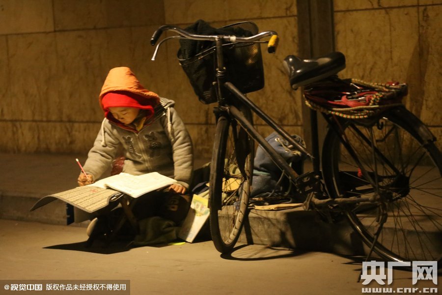 鄭州中原路京廣鐵路大橋下涵洞的馬路牙子邊，一位戴著紅色辣椒帽的小男孩趴在一個(gè)小馬扎上，上面鋪著一張車(chē)墊，在微弱的燈光下寫(xiě)作業(yè)