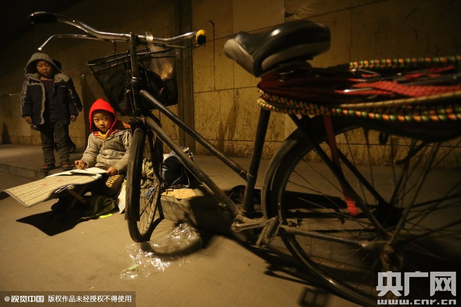 2015年11月8日晚，鄭州，7歲男孩涵洞橋下陪父母練攤，路邊燈下坐小馬扎上寫(xiě)作業(yè)。